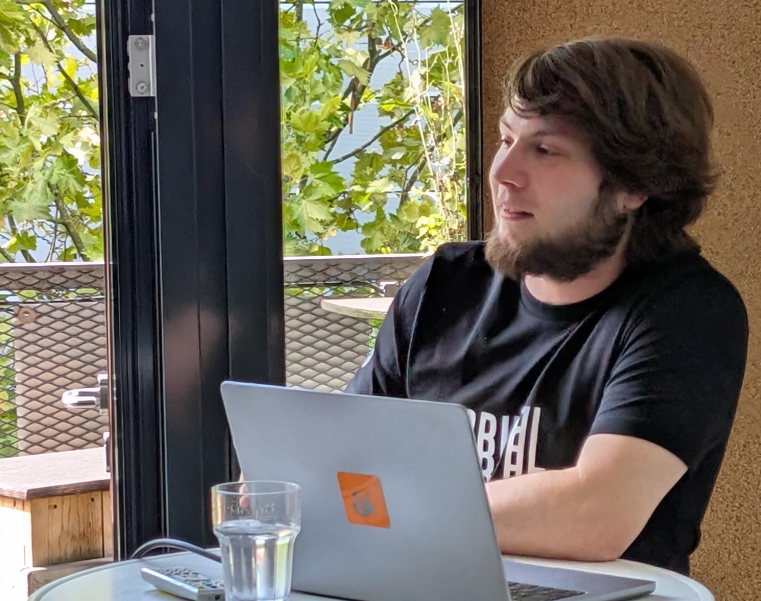 Ein junger Mann mit Bart und dunklem T-Shirt sitzt mit einem Laptop an einem runden Tisch in einem lichtdurchfluteten Raum.