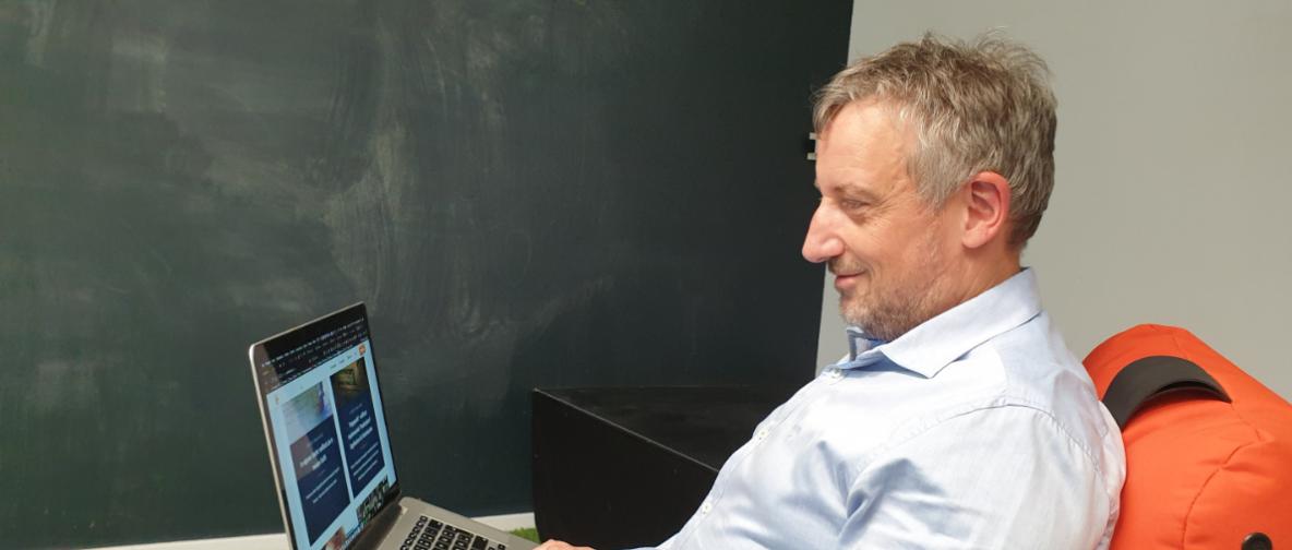 Ein Mann sitzt vor einer Tafel mit einem Laptop auf dem Schoß.