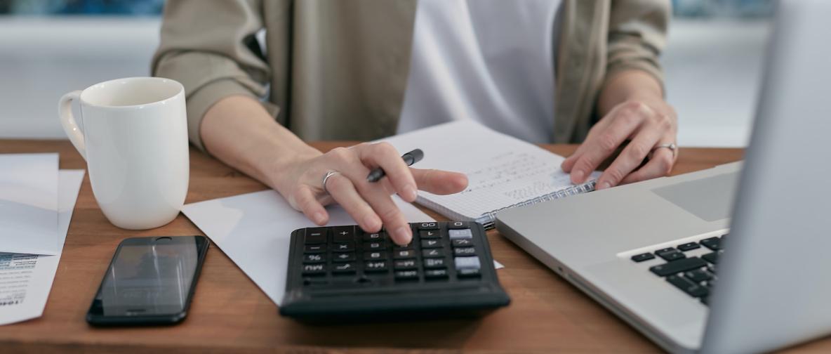 Person am Scheibtisch mit Notebook und Tischrechner