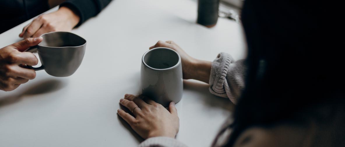 zwei Menschen sitzen sich fuer ein One-on-One Meeting an einem Tisch gegenüber. Jeweils eine Kaffeetassse in der Hand. Leicht von oben schraeg aufgenommen, so das im wesentliche nur die Unterarme samt Tassen zu sehen sind.