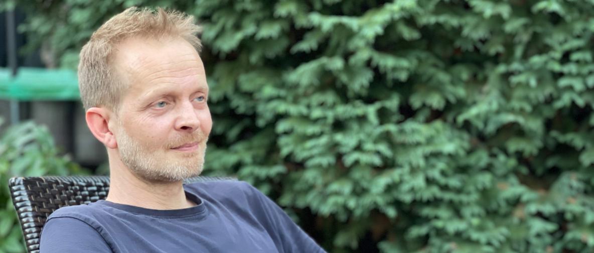 Mann mit kurzen blonden Haaren im Liegestuhl sitzend vor einem Baum. Kopf und Brustbereich sichtbar.