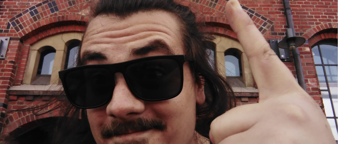 Alt-Text: Ein Mann mit langem Haar und Sonnenbrille macht ein Selfie vor einem Gebäude mit der Aufschrift "KAFFEERÖSTEREI". Er zeigt mit seinem Finger auf das Schild und lächelt leicht. Das Gebäude besteht aus roten Ziegelsteinen mit bogenförmigen Fenstern.