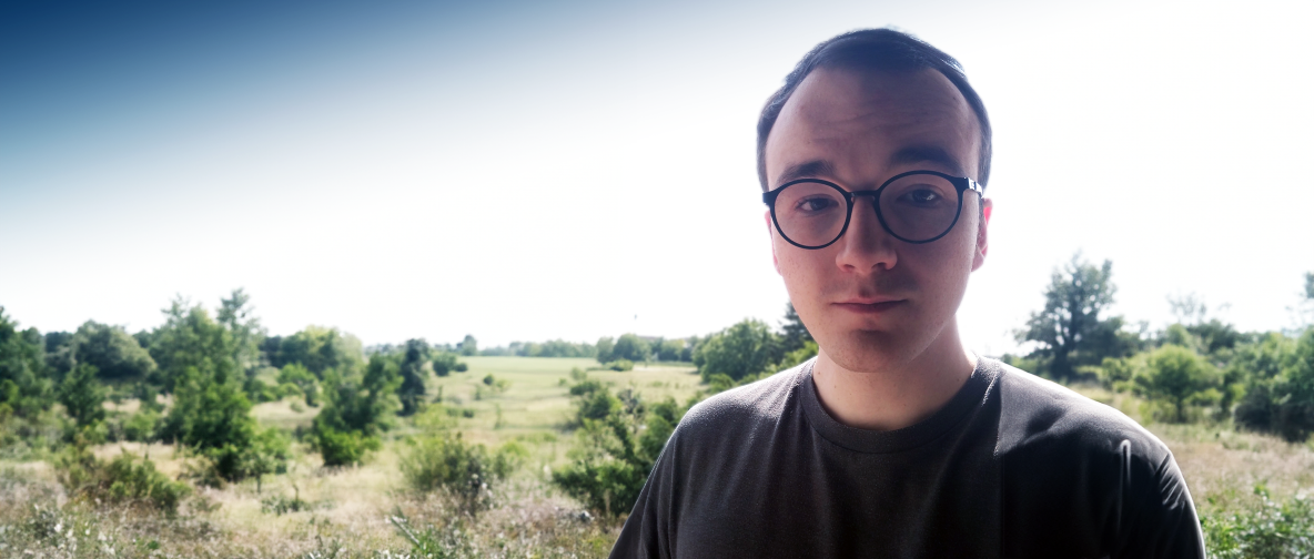 ### Bildbeschreibung Das Foto zeigt einen jungen Mann mit kurzen, dunklen Haaren und einer Brille mit runden Gläsern. Er trägt ein graues T-Shirt und steht im Freien. Im Hintergrund erstreckt sich eine weite Landschaft mit grünem, bewachsenen Grasland und vereinzelten Bäumen. Der Himmel ist klar und hell, was auf einen sonnigen Tag hinweist. Der Mann schaut direkt in die Kamera und lächelt leicht. ### ALT-Text Porträt eines jungen Mannes mit Brille im Freien vor einer grünen Landschaft mit Bäumen und kla