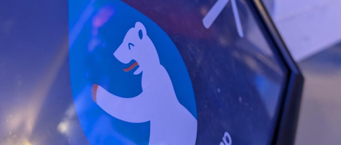 Ein Nahaufnahmebild zeigt das Logo des Drupal Camps Berlin mit einem weiĂen BĂ€ren in einem blauen Tropfensymbol. Der BĂ€r hat orangefarbene Akzente an Pfoten und Zunge. Die Szene ist in blauen und violetten Tönen gehalten.