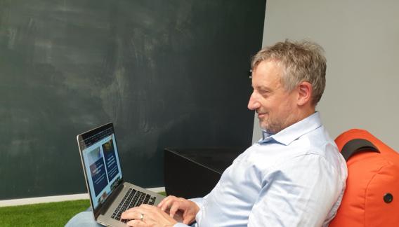 Ein Mann sitzt vor einer Tafel mit einem Laptop auf dem Schoß.