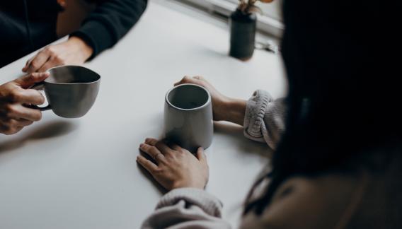 zwei Menschen sitzen sich fuer ein One-on-One Meeting an einem Tisch gegenüber. Jeweils eine Kaffeetassse in der Hand. Leicht von oben schraeg aufgenommen, so das im wesentliche nur die Unterarme samt Tassen zu sehen sind.