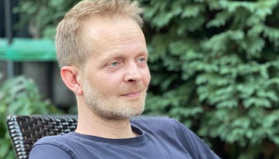 Mann mit kurzen blonden Haaren im Liegestuhl sitzend vor einem Baum. Kopf und Brustbereich sichtbar.