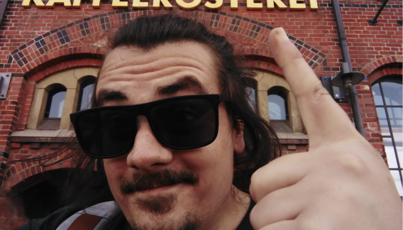 Alt-Text: Ein Mann mit langem Haar und Sonnenbrille macht ein Selfie vor einem Gebäude mit der Aufschrift "KAFFEERÖSTEREI". Er zeigt mit seinem Finger auf das Schild und lächelt leicht. Das Gebäude besteht aus roten Ziegelsteinen mit bogenförmigen Fenstern.
