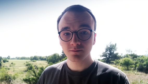 ### Bildbeschreibung  Das Foto zeigt einen jungen Mann mit kurzen, dunklen Haaren und einer Brille mit runden Gläsern. Er trägt ein graues T-Shirt und steht im Freien. Im Hintergrund erstreckt sich eine weite Landschaft mit grünem, bewachsenen Grasland und vereinzelten Bäumen. Der Himmel ist klar und hell, was auf einen sonnigen Tag hinweist. Der Mann schaut direkt in die Kamera und lächelt leicht.  ### ALT-Text  Porträt eines jungen Mannes mit Brille im Freien vor einer grünen Landschaft mit Bäumen und kla