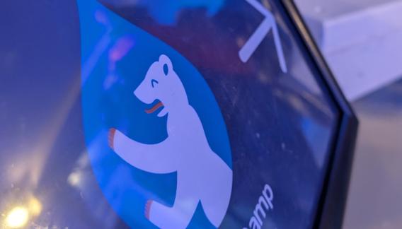 Ein Nahaufnahmebild zeigt das Logo des Drupal Camps Berlin mit einem weißen Bären in einem blauen Tropfensymbol. Der Bär hat orangefarbene Akzente an Pfoten und Zunge. Die Szene ist in blauen und violetten Tönen gehalten.