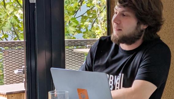 Ein junger Mann mit Bart und dunklem T-Shirt sitzt mit einem Laptop an einem runden Tisch in einem lichtdurchfluteten Raum.