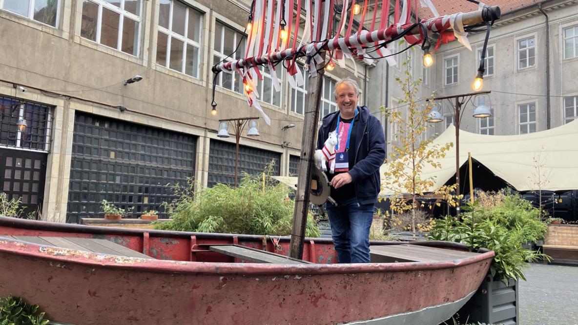 Ein Mann steht in einem dekorativen roten Boot in einem Innenhof, das mit rot-weiß gestreiften Stoffen geschmückt ist. Er hält ein Plüschpferd mit einem rot-weiß gestreiften Schal in der Hand und lächelt in die Kamera. Um das Boot herum sind Pflanzen und Lichter zu sehen, die eine gemütliche Atmosphäre schaffen.