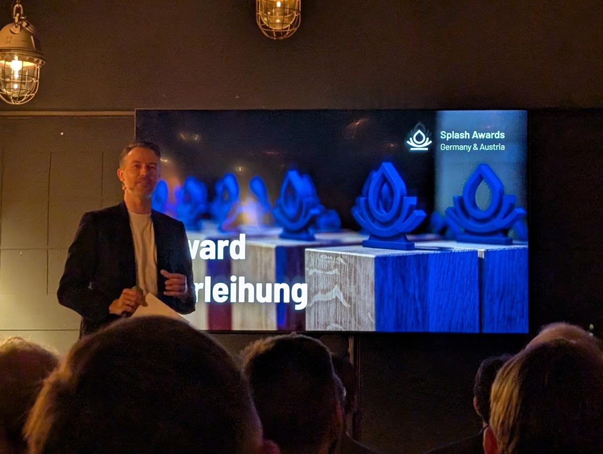 Moderator bei der Preisverleihung der Splash Awards 2024 in Deutschland, mit den Trophäen für ausgezeichnete Drupal-Projekte im Hintergrund auf der Leinwand.