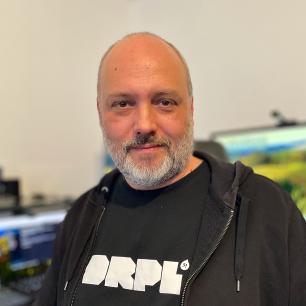 Ein Mann mit kurzem grauem Bart und rasiertem Kopf trägt ein schwarzes T-Shirt mit der Aufschrift „DRPL“ und eine schwarze Kapuzenjacke. Er blickt freundlich in die Kamera. Im Hintergrund sind mehrere Bildschirme und ein Schreibtisch mit Technik-Equipment zu sehen.
