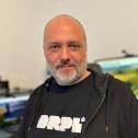Ein Mann mit kurzem grauem Bart und rasiertem Kopf trägt ein schwarzes T-Shirt mit der Aufschrift „DRPL“ und eine schwarze Kapuzenjacke. Er blickt freundlich in die Kamera. Im Hintergrund sind mehrere Bildschirme und ein Schreibtisch mit Technik-Equipment zu sehen.