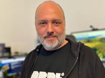 Ein Mann mit kurzem grauem Bart und rasiertem Kopf trägt ein schwarzes T-Shirt mit der Aufschrift „DRPL“ und eine schwarze Kapuzenjacke. Er blickt freundlich in die Kamera. Im Hintergrund sind mehrere Bildschirme und ein Schreibtisch mit Technik-Equipment zu sehen.